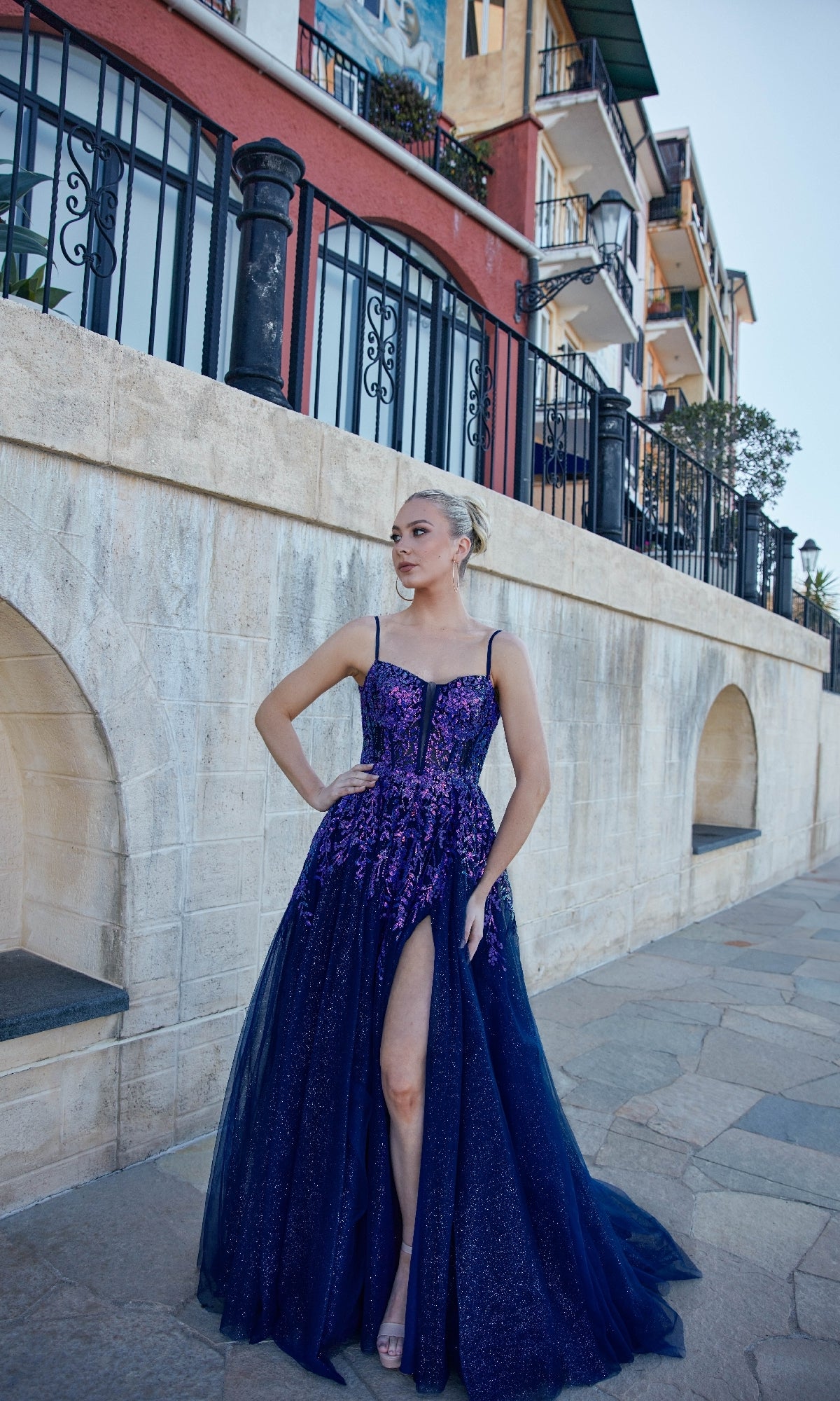 A woman in a purple glitter tulle long formal dress, Tina Holly TE216B, with a high-leg slit stands on a stone walkway before a building with balconies and arched walls.